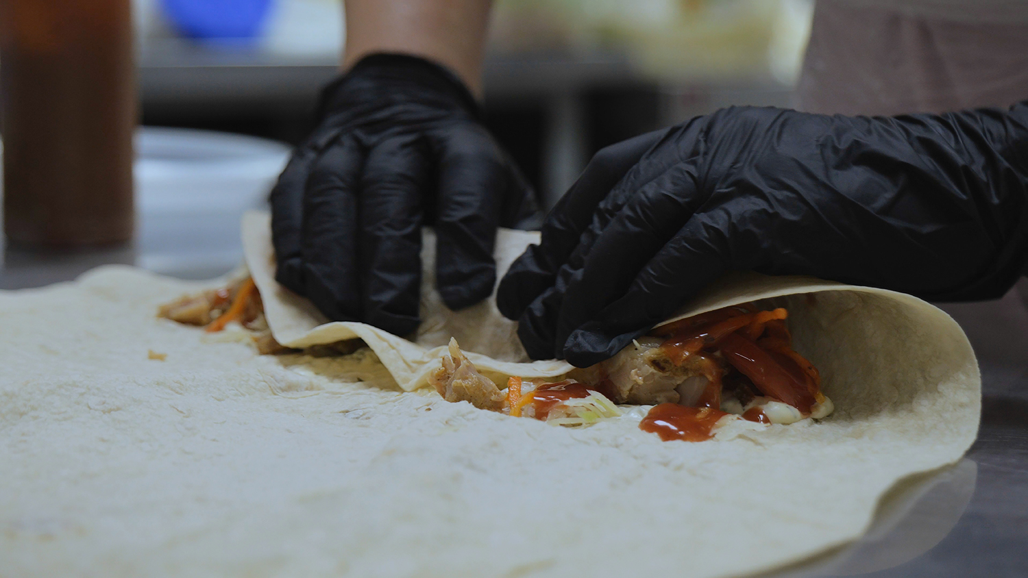 Chef in black gloves hand-rolling a fresh wrap with chicken, vegetables, and chipotle sauce.